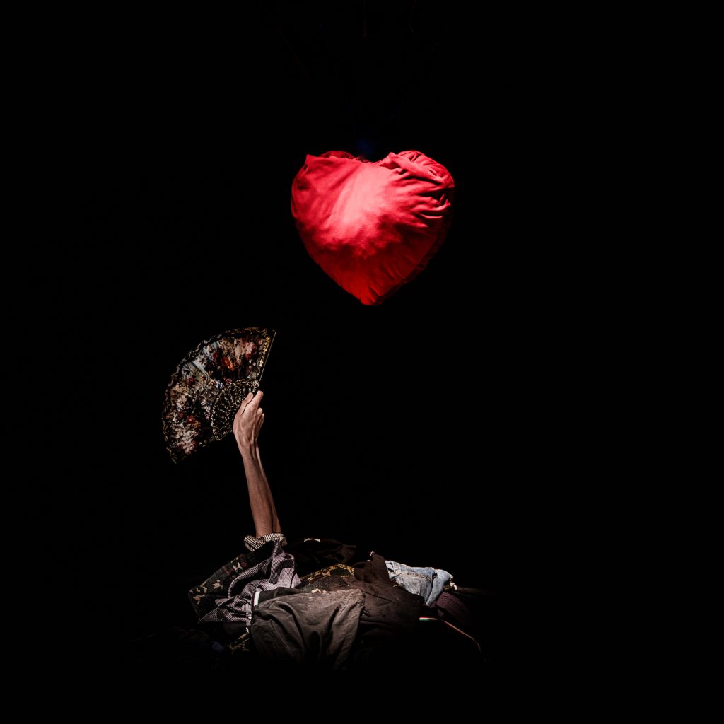 Immagine di mano che tiene un ventaglio aperto vicino a un cuore rosso di peluche appeso su una struttura sospesa. La foto è di Giorgio Meneghetti e ritrae un momento dello spettacolo La Voce di Carola Minincleri Colussi / Farmacia Zooè