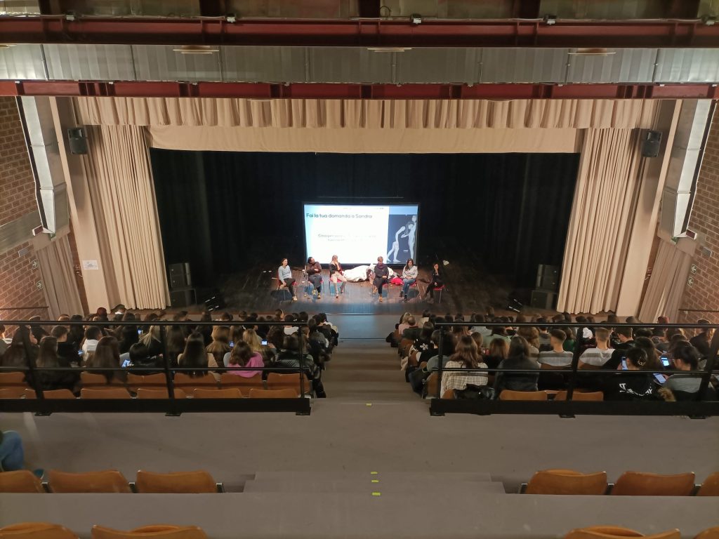 Immagine della gradinata del Teatro del Parco di Mestre (YouTheater) con giovani seduti di spalle. Ascoltano 6 persone che parlano davanti a uno schermo sul palco. La foto è tratta dall'evento "La Voce. Esprimi un desiderio"