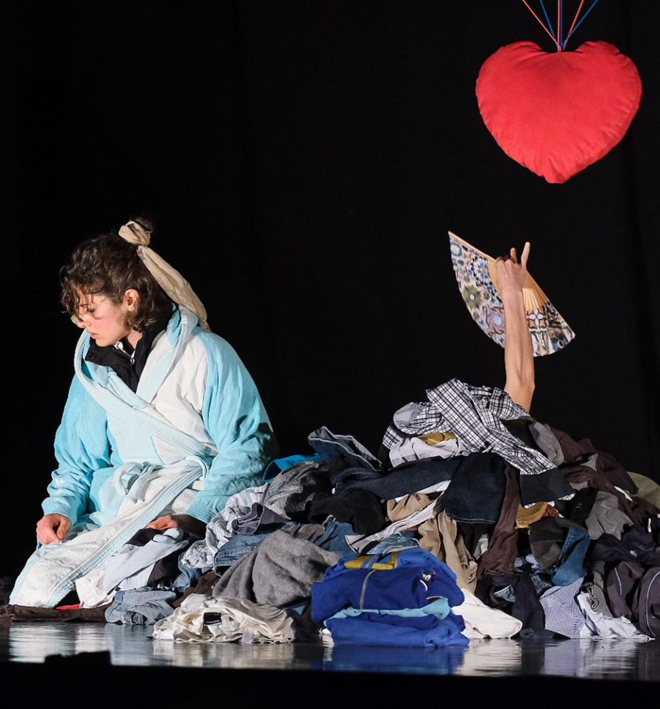 Donna inginocchiata vestita con molti strati di abiti dà le spalle a un cuore di pezza appeso e da un mucchio di vestiti esce una mano che stringe un ventaglio.